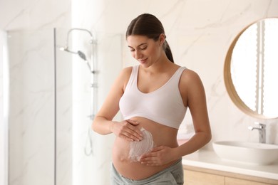 Young pregnant woman applying cosmetic product on belly in bathroom