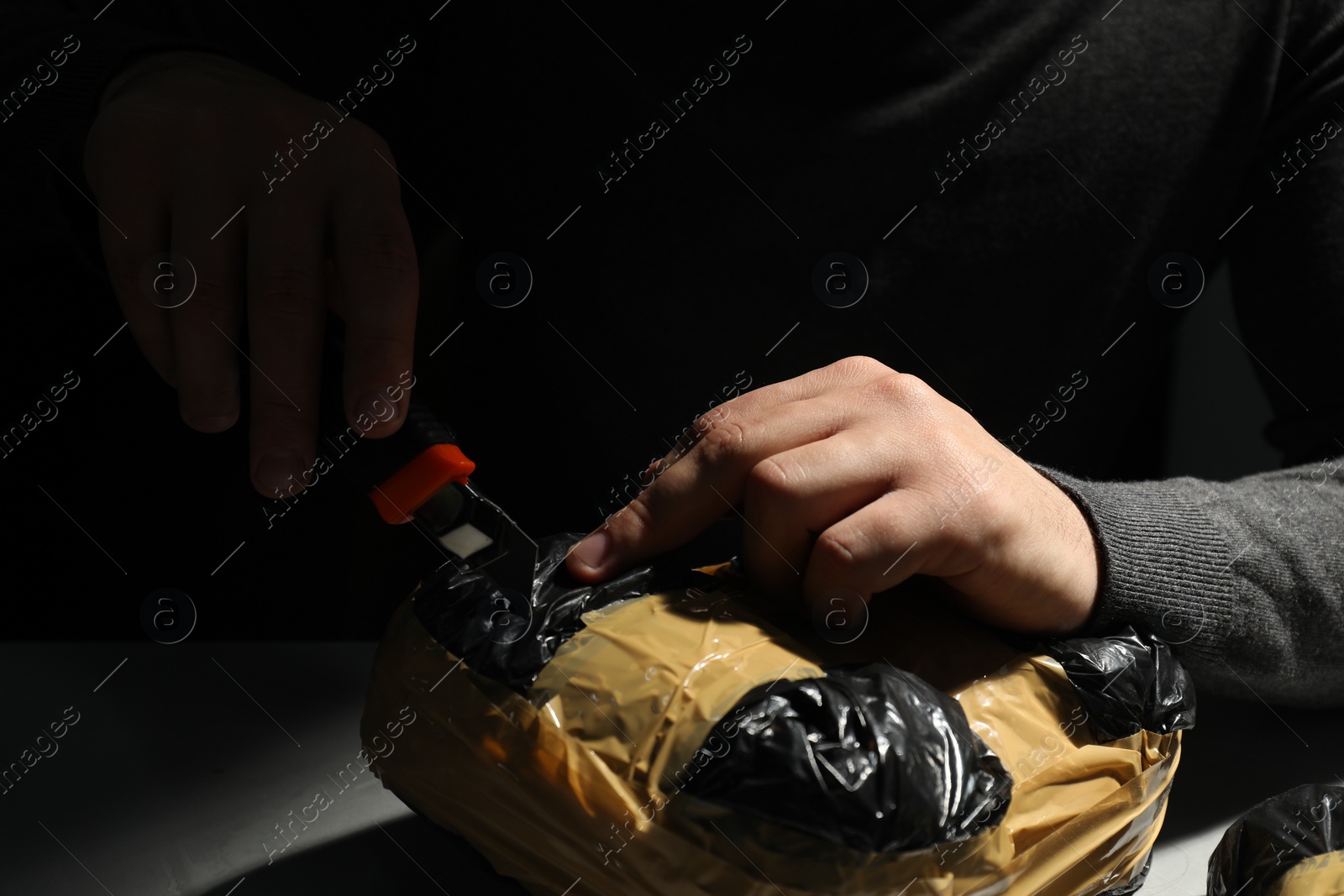 Photo of Smuggling and drug trafficking. Man opening package of narcotics with box cutter in darkness, closeup