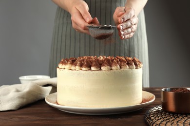 Photo of Woman dusting delicious tiramisu cake with cocoa powder at wooden table, closeup