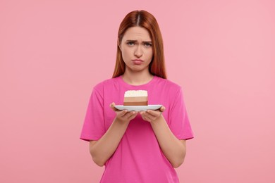 Unhappy young woman with piece of tasty cake on pink background