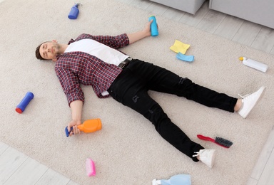 Photo of Tired man lying on carpet surrounded by cleaning supplies