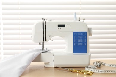 Photo of White sewing machine, scissors, measuring tape and fabric on wooden table indoors