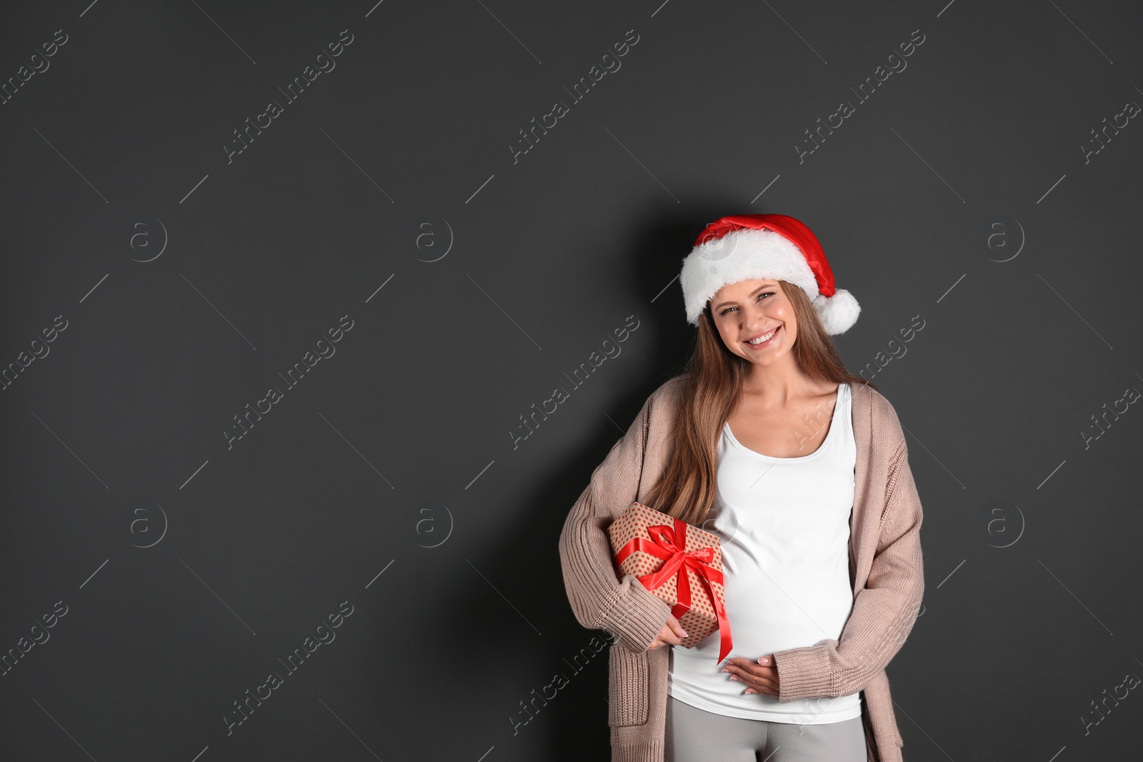 Photo of Young pregnant woman in Santa hat with Christmas gift on grey background, space for text