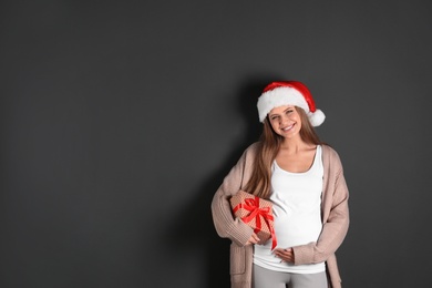 Photo of Young pregnant woman in Santa hat with Christmas gift on grey background, space for text