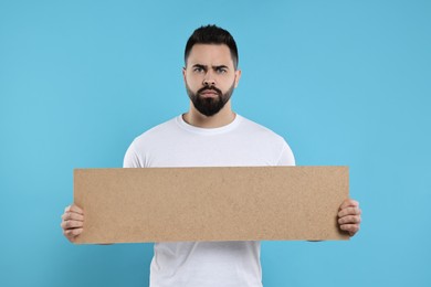 Upset man holding blank cardboard banner on light blue background, space for text