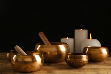 Golden singing bowls with mallets and burning candles on wooden table against dark background
