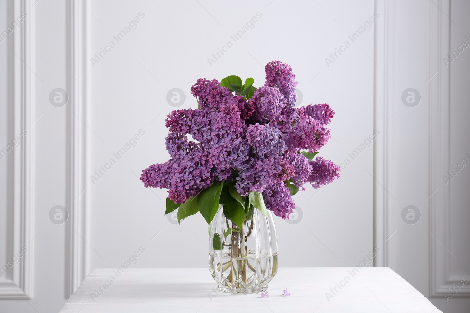 Photo of Beautiful lilac flowers in vase on white table near wall