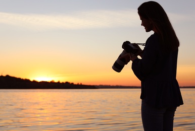 Young female photographer holding professional camera on riverside at sunset