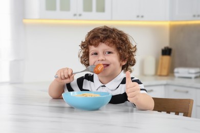 Cute little boy eating sausage and showing thumbs up at table in kitchen