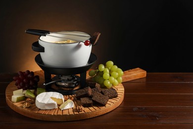 Forks with pieces of grape, bread, fondue pot with melted cheese and snacks on wooden table. Space for text