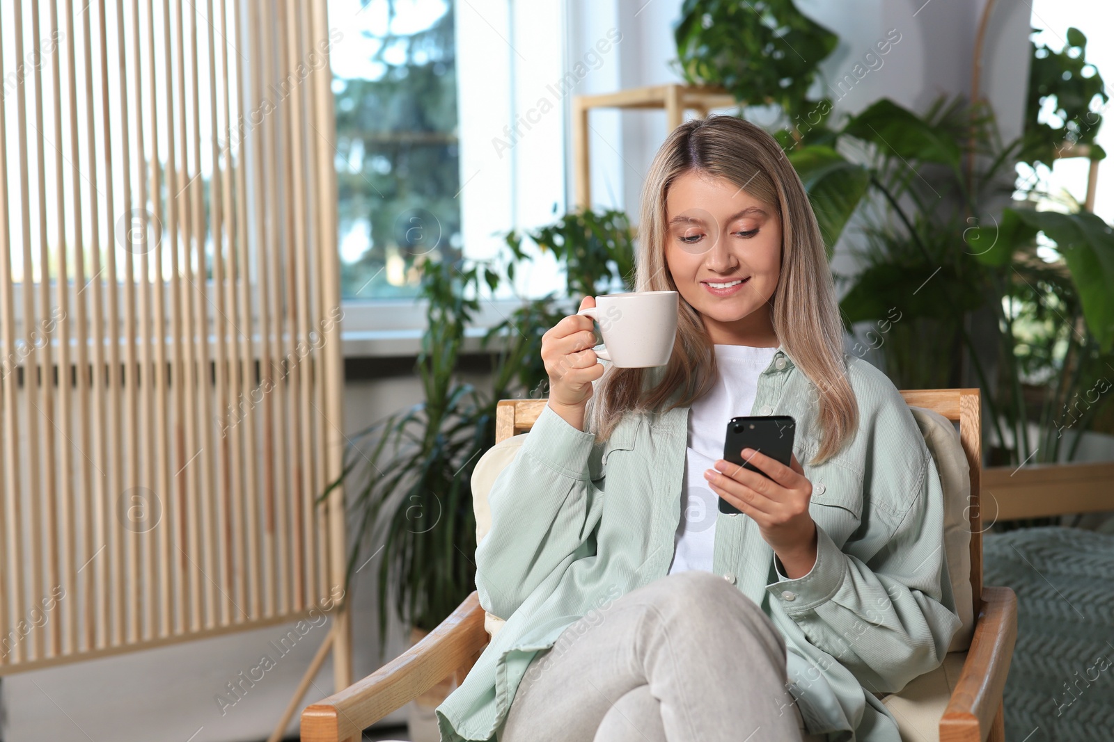 Photo of Woman with smartphone and cup of drink sitting in armchair at home