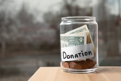 Glass jar with money and label DONATION on table against blurred background. Space for text