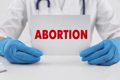 Doctor holding paper card with word Abortion at white table, closeup