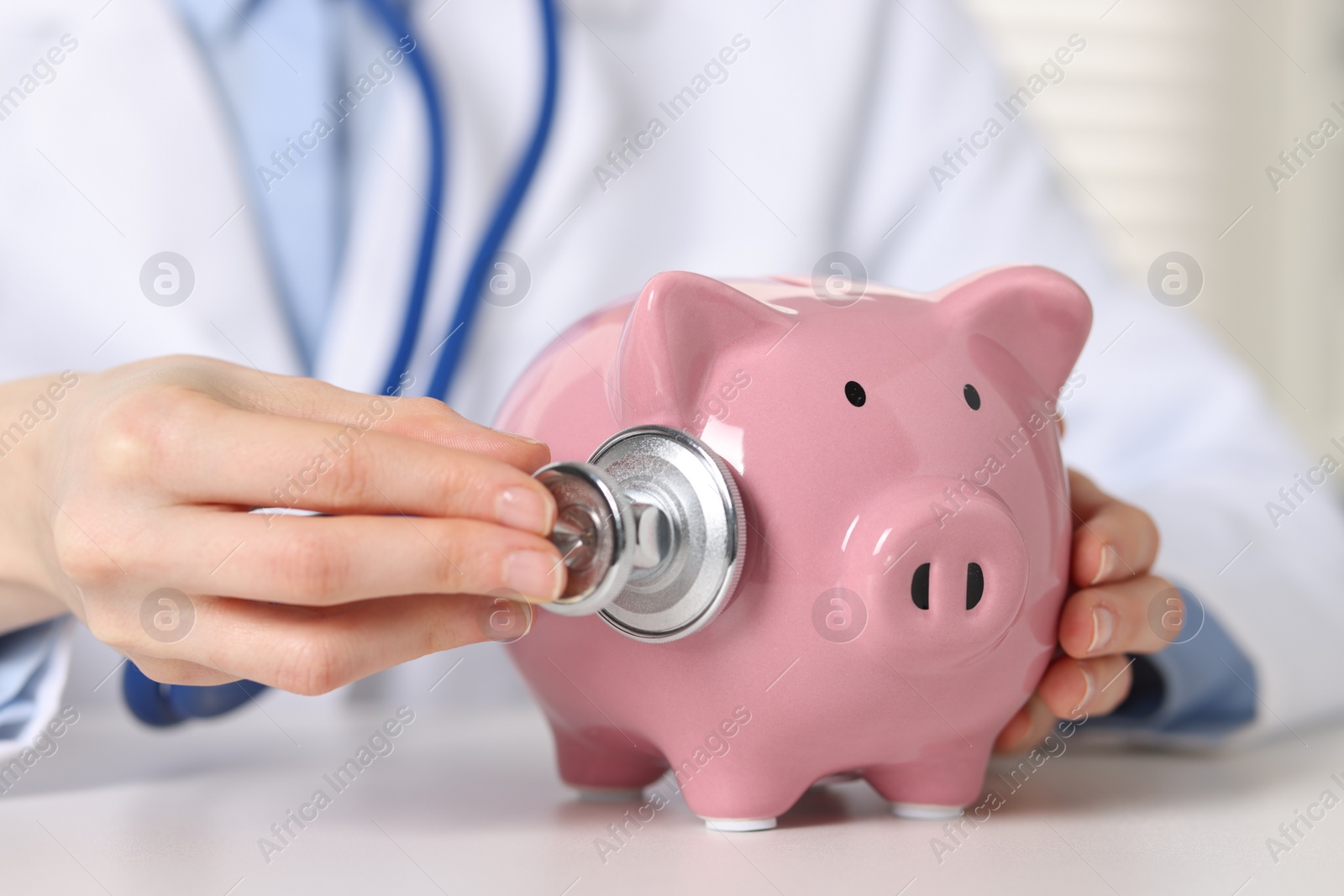 Photo of Doctor with piggy bank at white table indoors, closeup