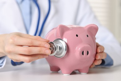 Doctor with piggy bank at white table indoors, closeup