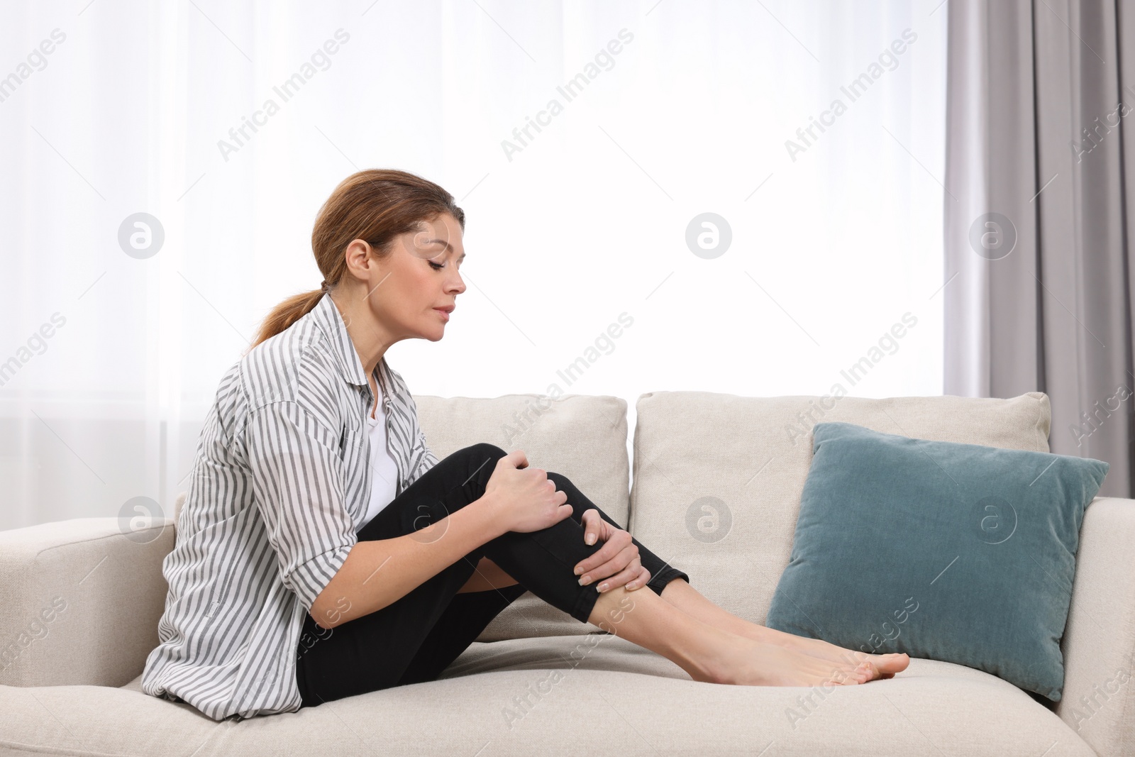 Photo of Woman suffering from leg pain on sofa indoors. Arthritis symptoms