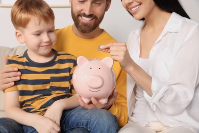 Photo of Happy family putting coin into piggy bank indoors, closeup