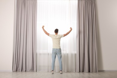 Photo of Man stretching near window with beautiful curtains at home, back view