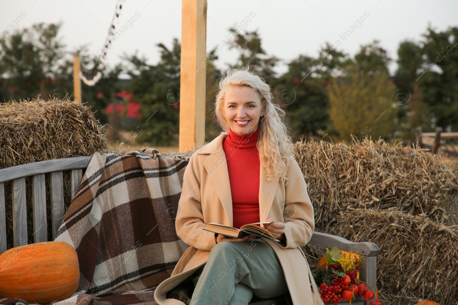 Photo of Beautiful woman with book sitting on wooden bench near pumpkins and hay bales outdoors. Autumn season