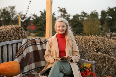 Photo of Beautiful woman with book sitting on wooden bench near pumpkins and hay bales outdoors. Autumn season