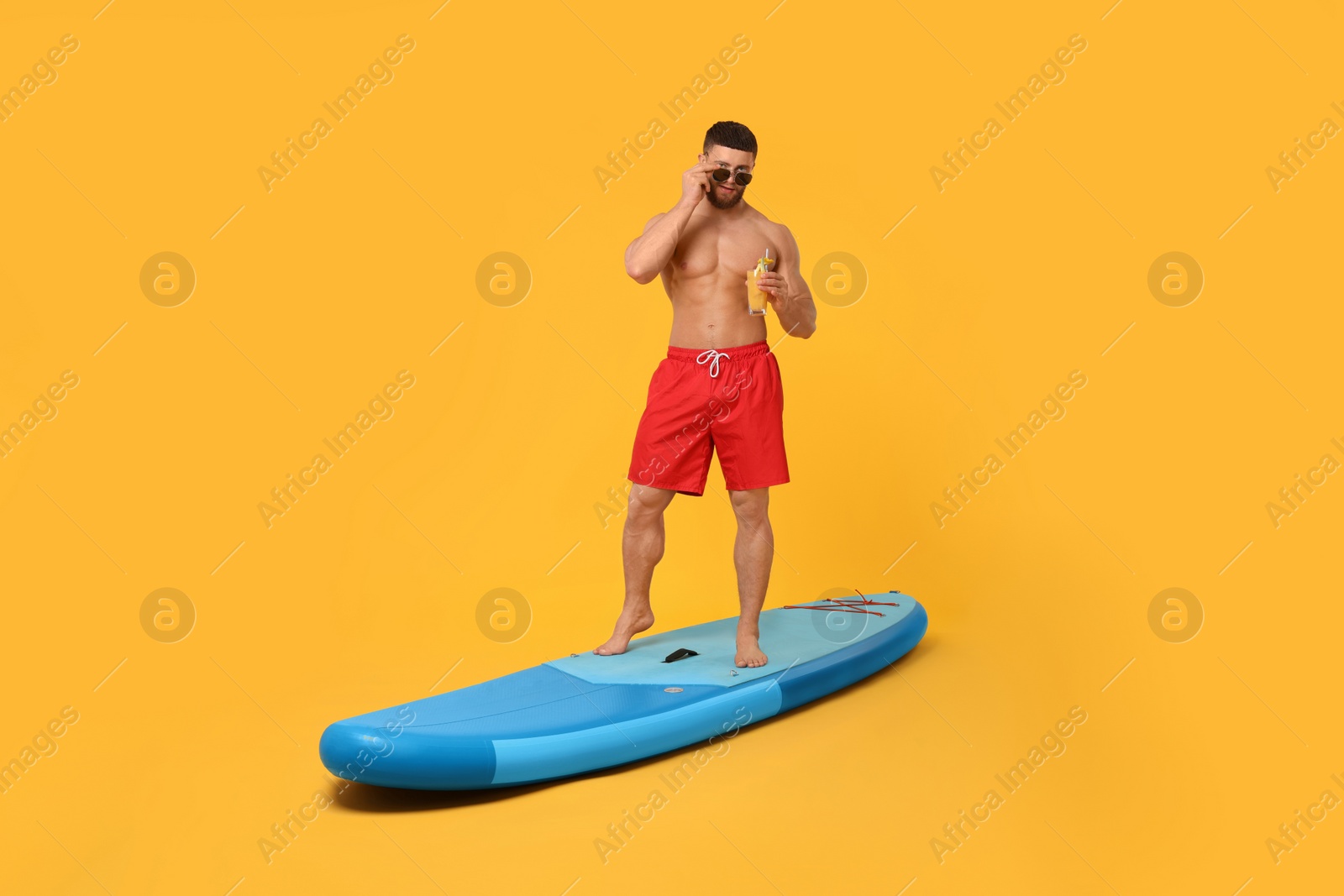 Photo of Man with refreshing drink posing on SUP board against orange background