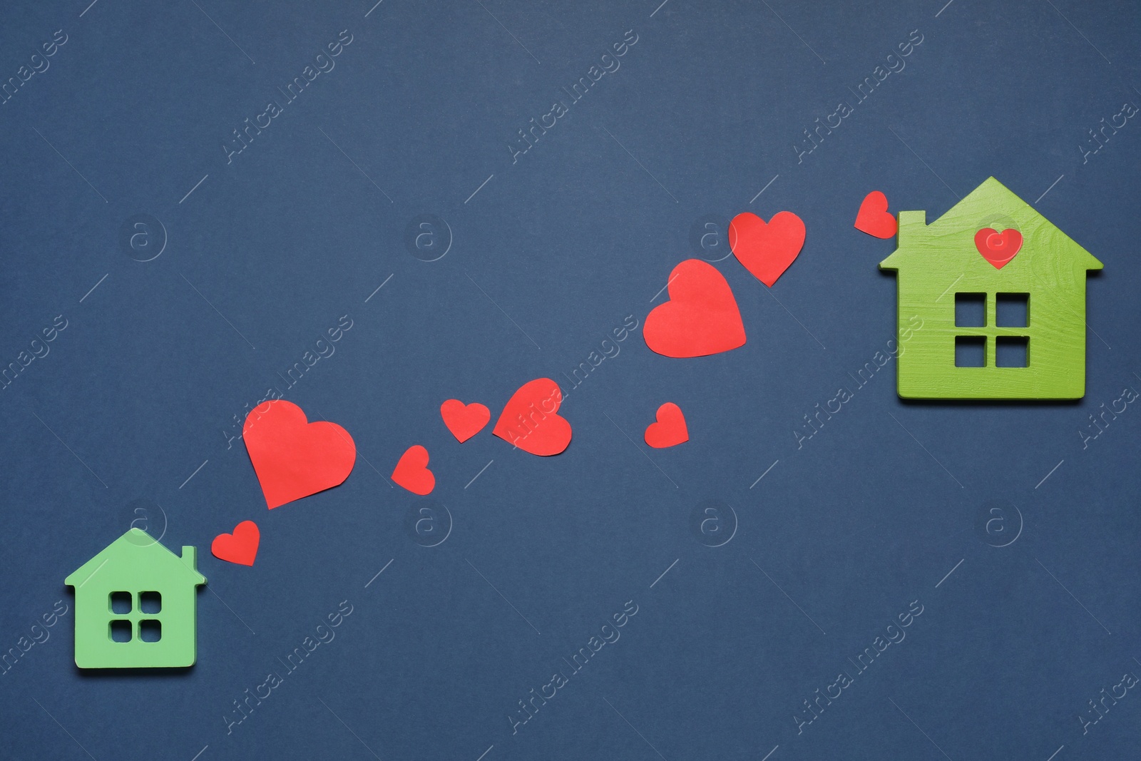 Photo of Decorative hearts between two green house models on blue background symbolizing connection in long-distance relationship, flat lay