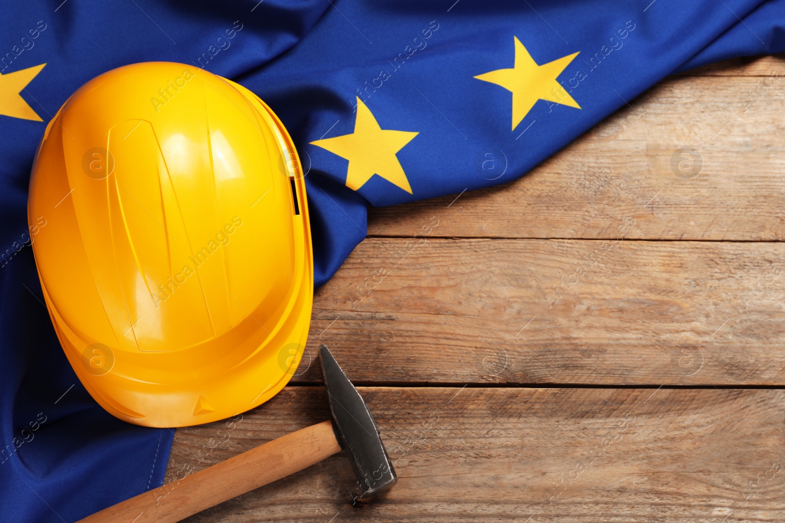 Photo of Yellow hard hat, hammer and flag of European Union on wooden table, flat lay. Space for text