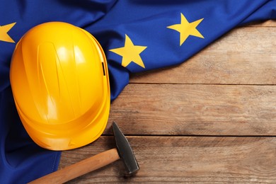 Photo of Yellow hard hat, hammer and flag of European Union on wooden table, flat lay. Space for text
