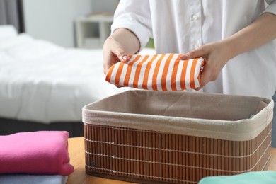 Woman putting folded clothes into storage basket at table indoors, closeup