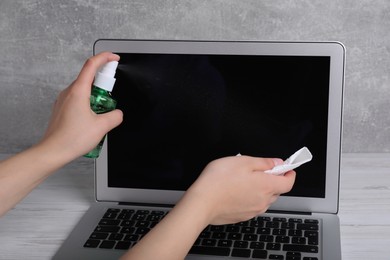 Photo of Woman cleaning laptop with sanitizer and paper towel at white wooden table, closeup