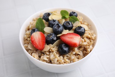 Photo of Tasty oatmeal with strawberries, blueberries and almond petals in bowl on white tiled table, closeup