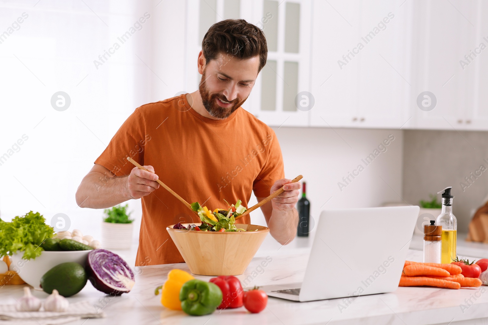 Photo of Man making dinner while watching online cooking course via laptop in kitchen