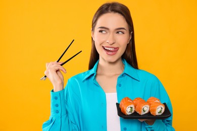 Photo of Happy young woman with plate of sushi rolls and chopsticks on orange background