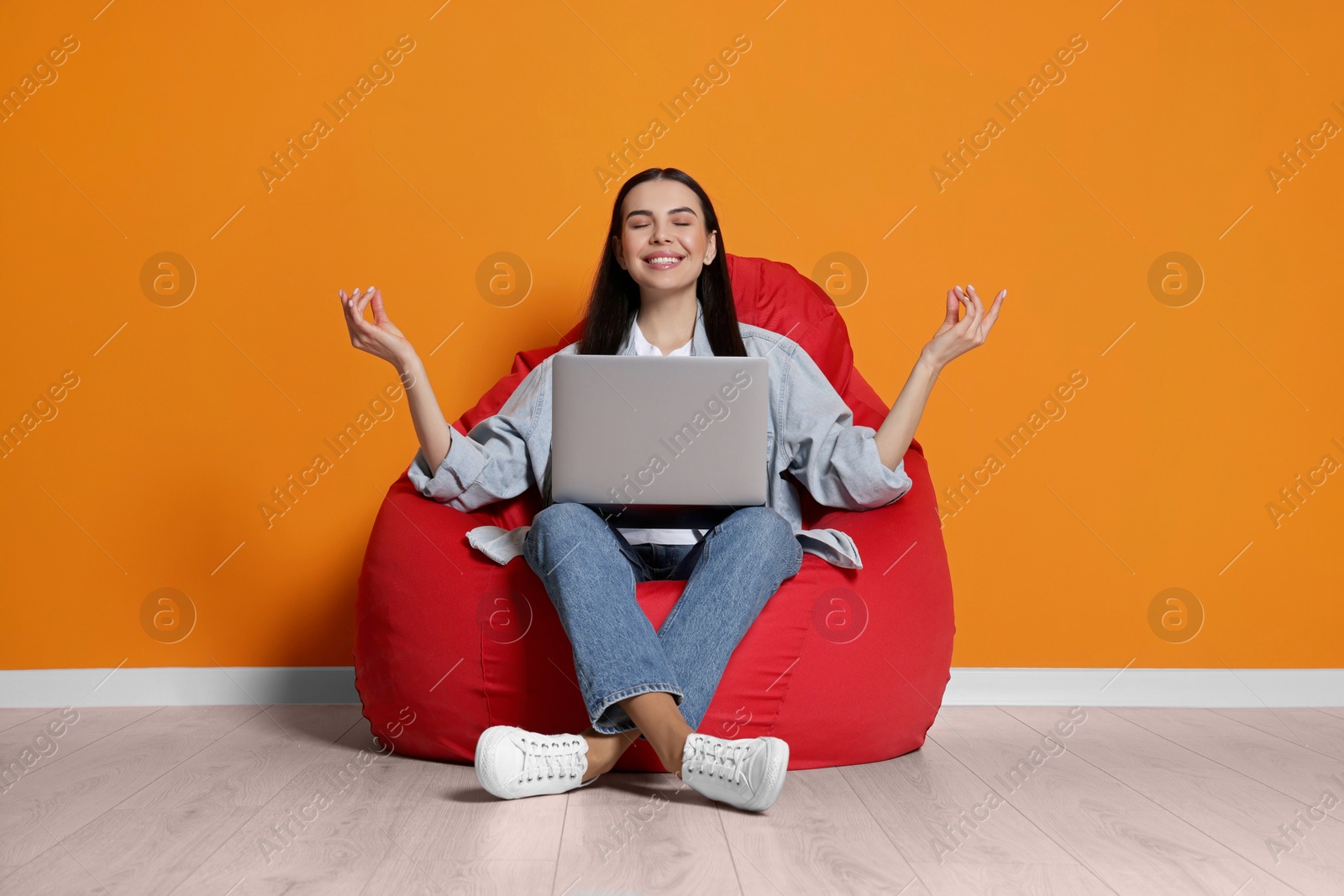 Photo of Happy woman with laptop sitting on beanbag chair and meditating near orange wall