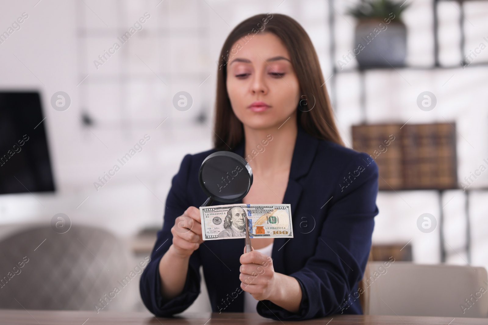 Photo of Expert authenticating 100 dollar banknote with magnifying glass at table in office, focus on hand. Fake money concept