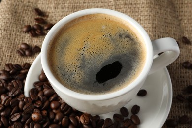 Photo of Cup of hot aromatic coffee and roasted beans on sackcloth, closeup
