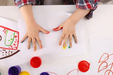 Little child making hand prints on paper with painted palms at white table, top view