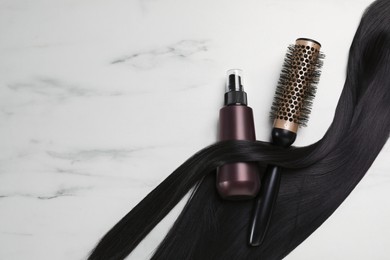 Spray bottle with thermal protection, lock of brunette hair and hairbrush on white marble table, flat lay. Space for text