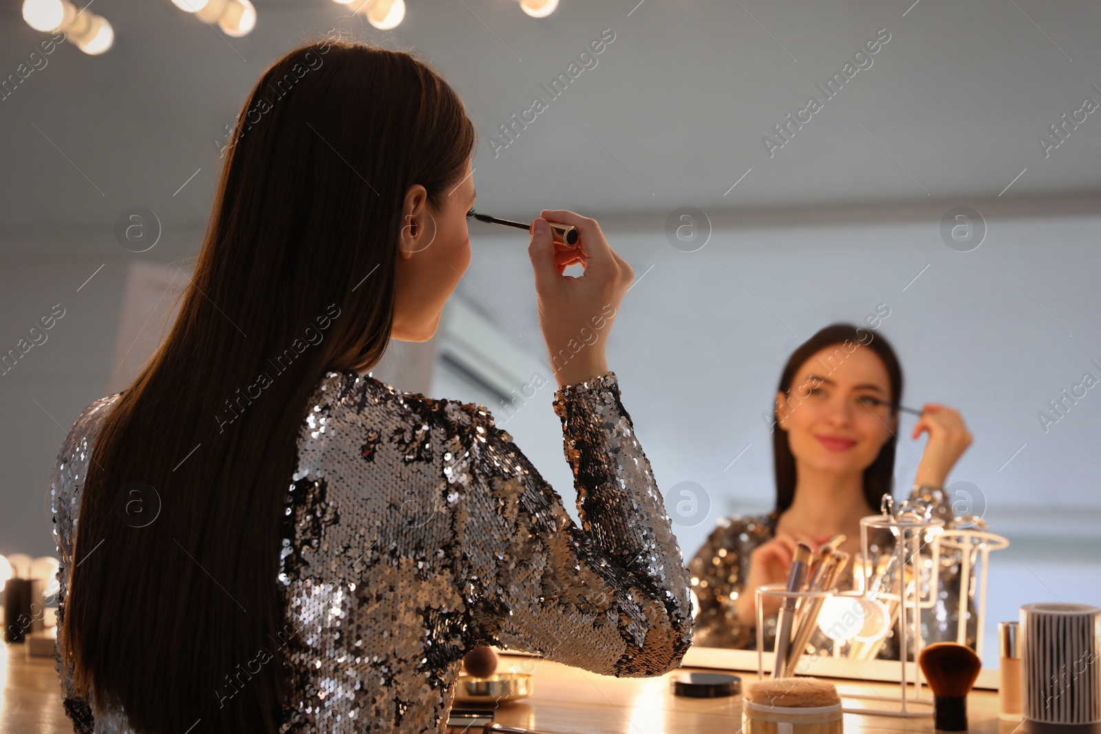 Photo of Young woman applying make up near illuminated mirror indoors