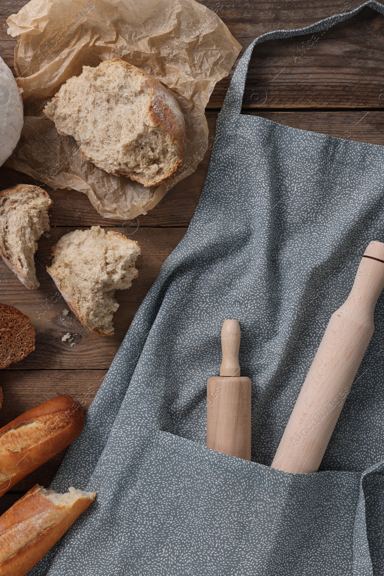 Photo of Clean kitchen apron with rolling pins and different types of bread on wooden table, flat lay