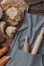 Photo of Clean kitchen apron with rolling pins and different types of bread on wooden table, flat lay