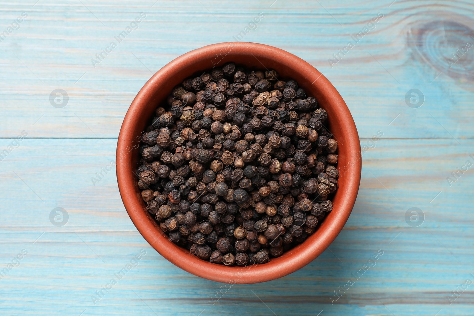 Photo of Aromatic spice. Black pepper in bowl on light blue wooden table, top view