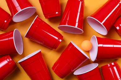 Photo of Plastic cups and ball for beer pong on orange background, flat lay