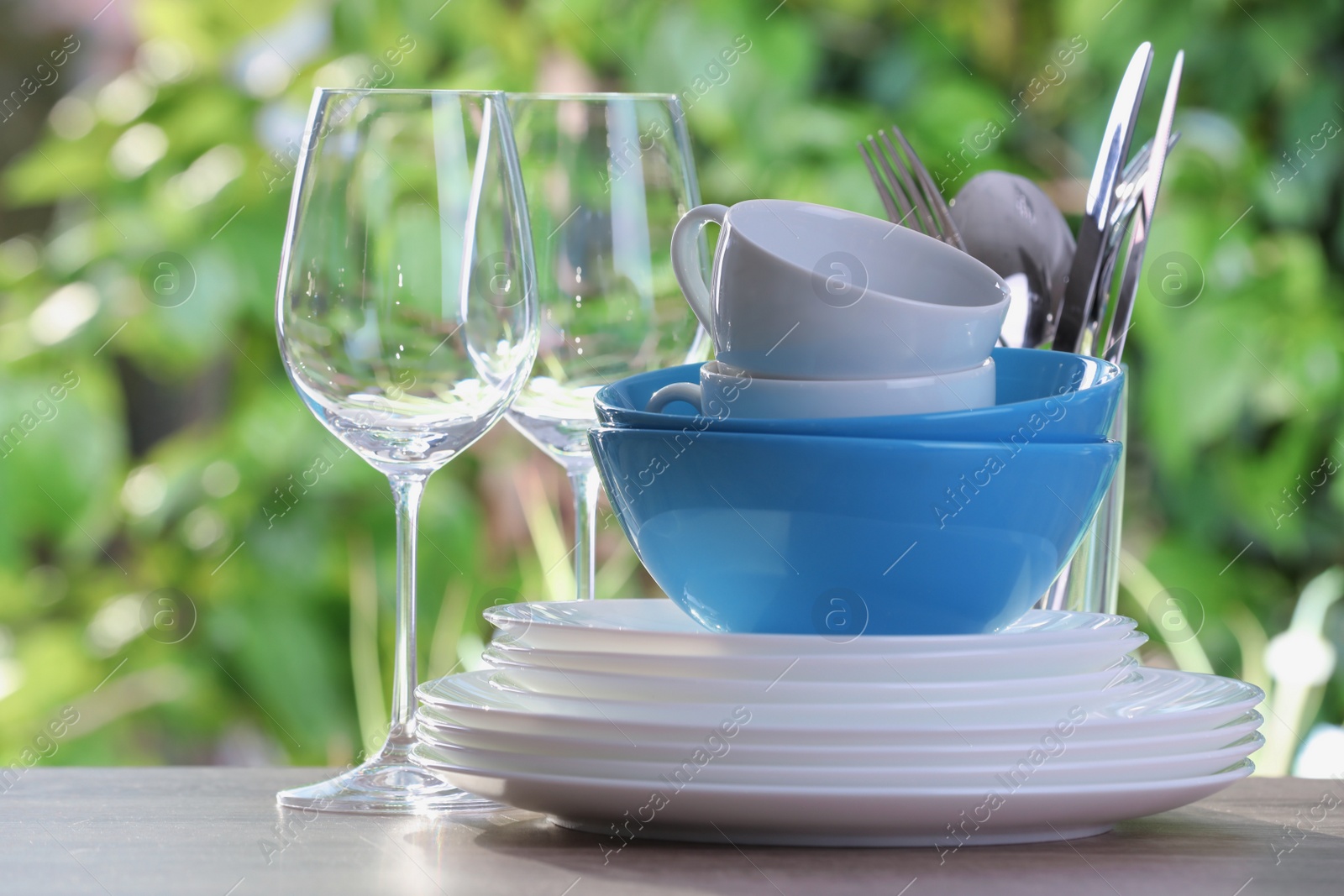 Photo of Clean dishes, cups, shiny cutlery and glasses on wooden table against blurred background