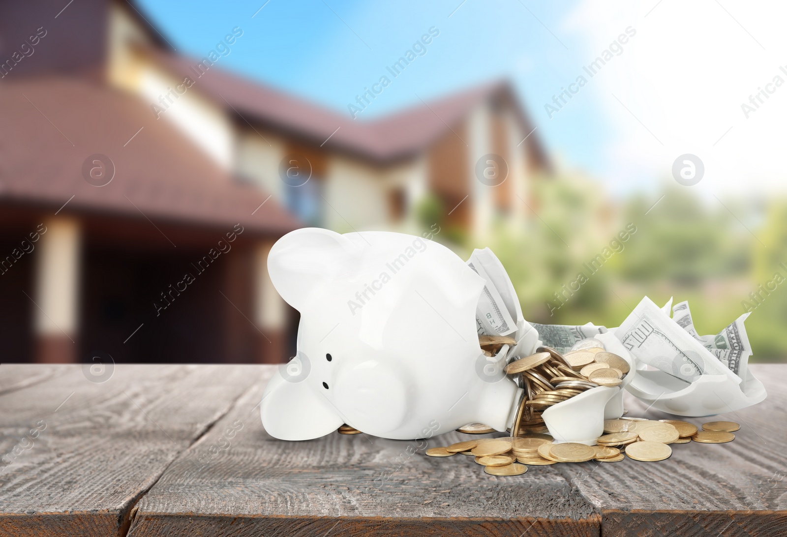 Image of Broken piggy bank with money on wooden surface and blurred view of beautiful house. Mortgage concept