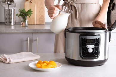 Woman pouring milk into modern multi cooker for making delicious yogurt at table, closeup