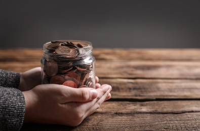 Photo of Woman holding donation jar with coins at wooden table, closeup. Space for text