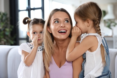 Photo of Cute little girls telling secrets to their mother in living room