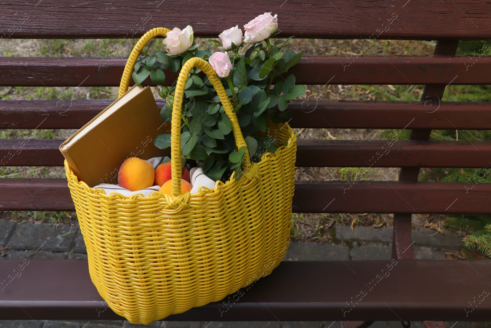 Photo of Yellow wicker bag with roses, book and peaches on bench outdoors