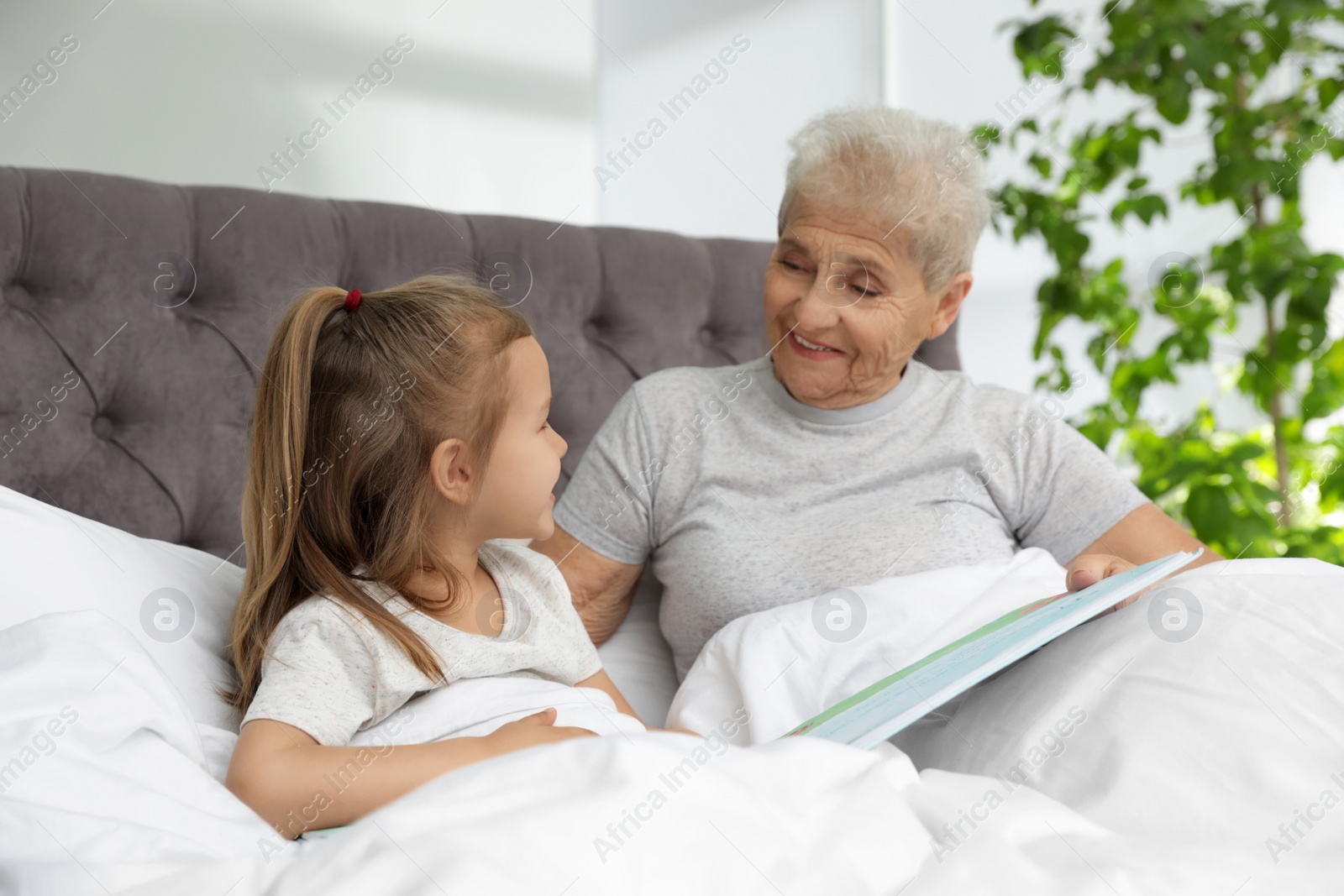 Photo of Cute girl and her grandmother reading book on bed at home